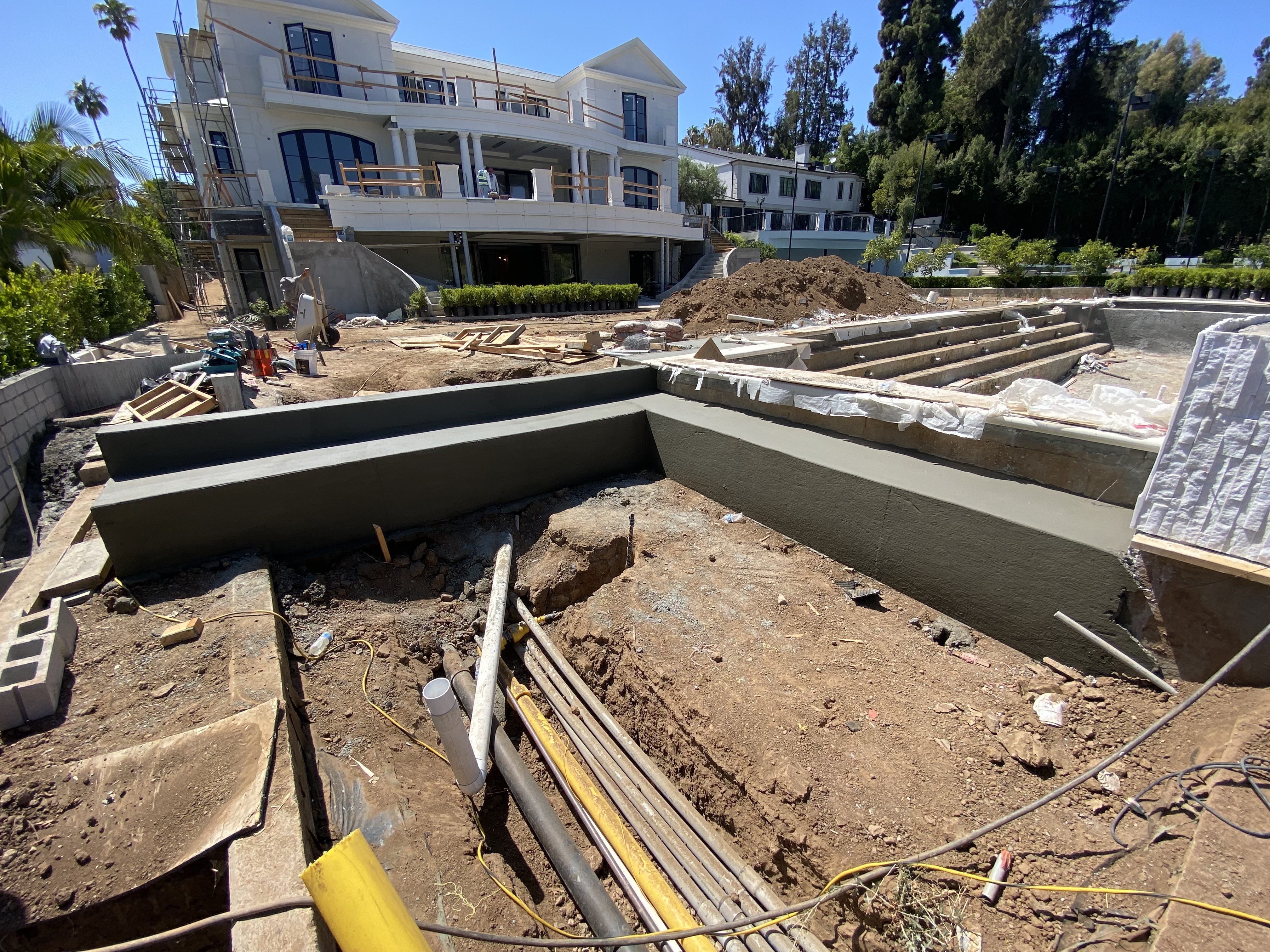 Image 8: The back of a 3 story house with the ground made of dirt.  There is the start of a pool on the right hand side and pipes exposed in the bottom center surrounded on 2 side with cement framing.